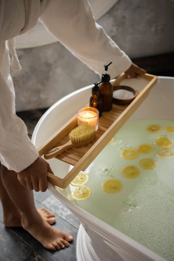 A bath with a woman's hand resting a tray containing a candle, brush, dispenser bottles and salt. Photo by Monstera Production via Pexels